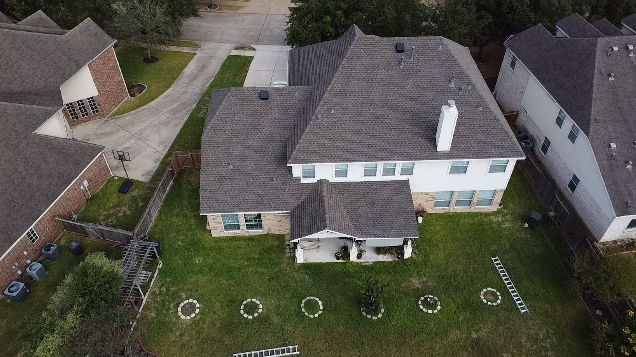 A drone-view shot of a suburban two-story house with a grey shingled roof, a chimney, and a grassy backyard.