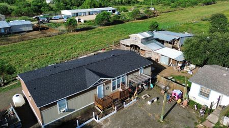 An aerial view of a single-story beige house with a dark roof, adjacent to outbuildings and a green field.