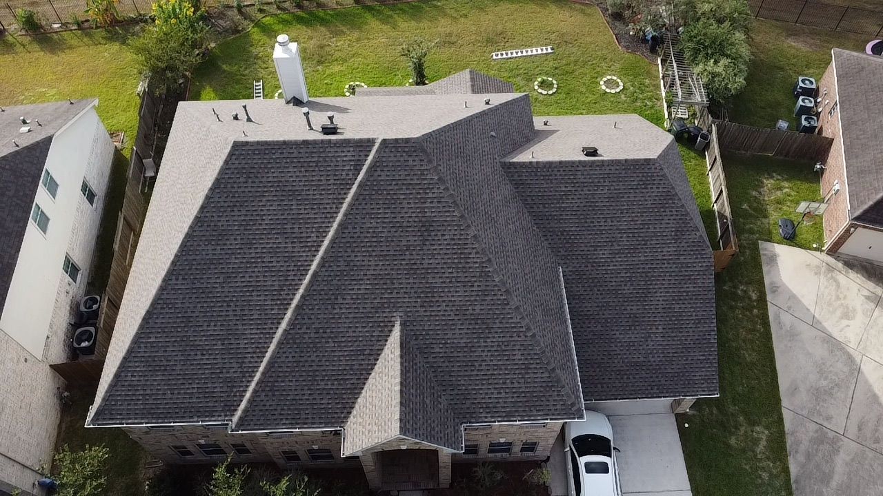 Aerial view of a residential shingle roof with a chimney and garage, surrounded by a lawn.