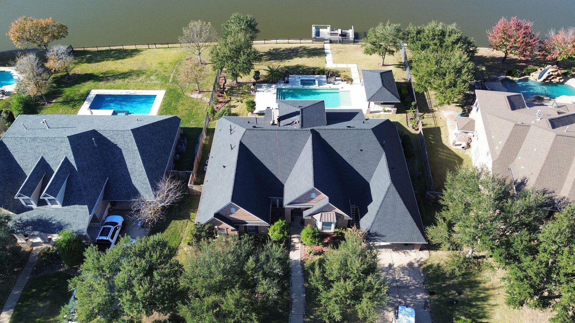An aerial view of three suburban homes with swimming pools near a lake, featuring dark gray shingled roofs and green yards.