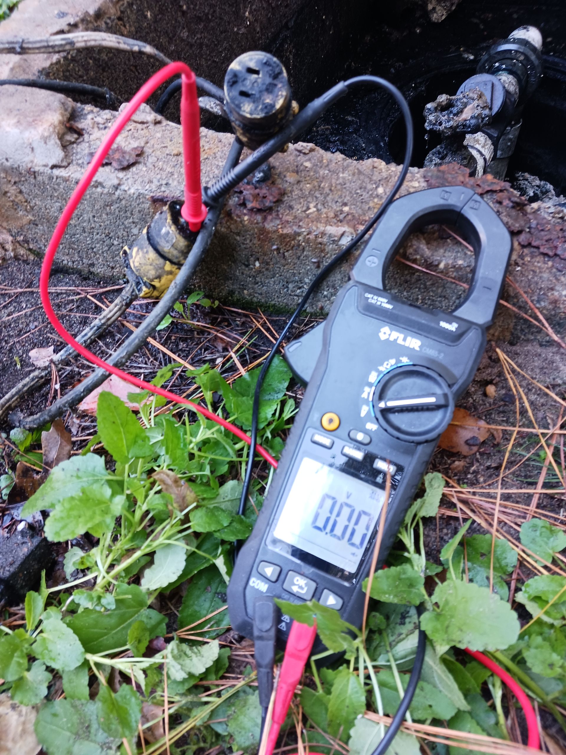 Clamp meter measuring electricity on a damaged outlet, with red and black leads connected. The meter reads 0.00.