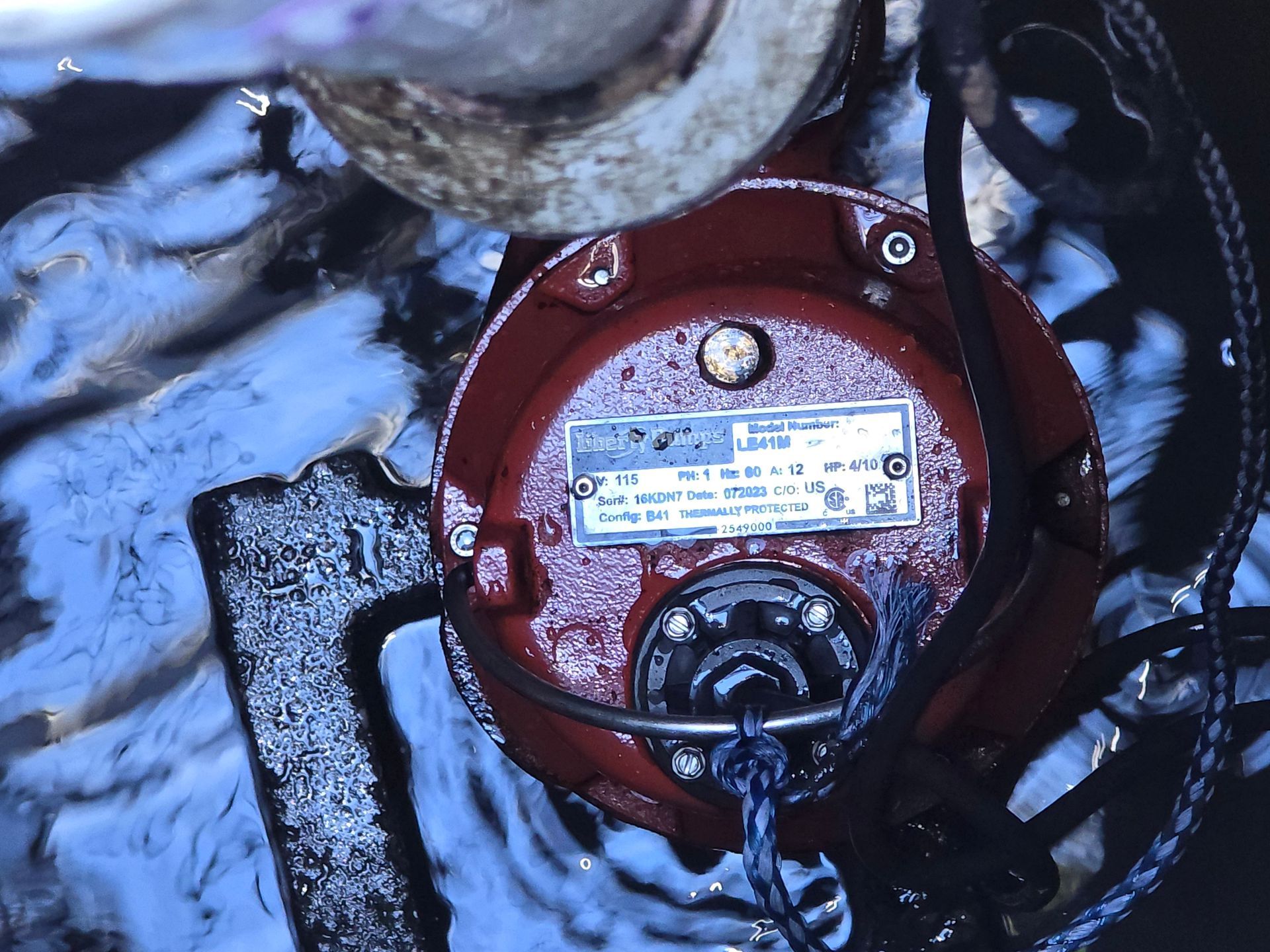 Red submersible pump in water, with attached cables and a metallic label.