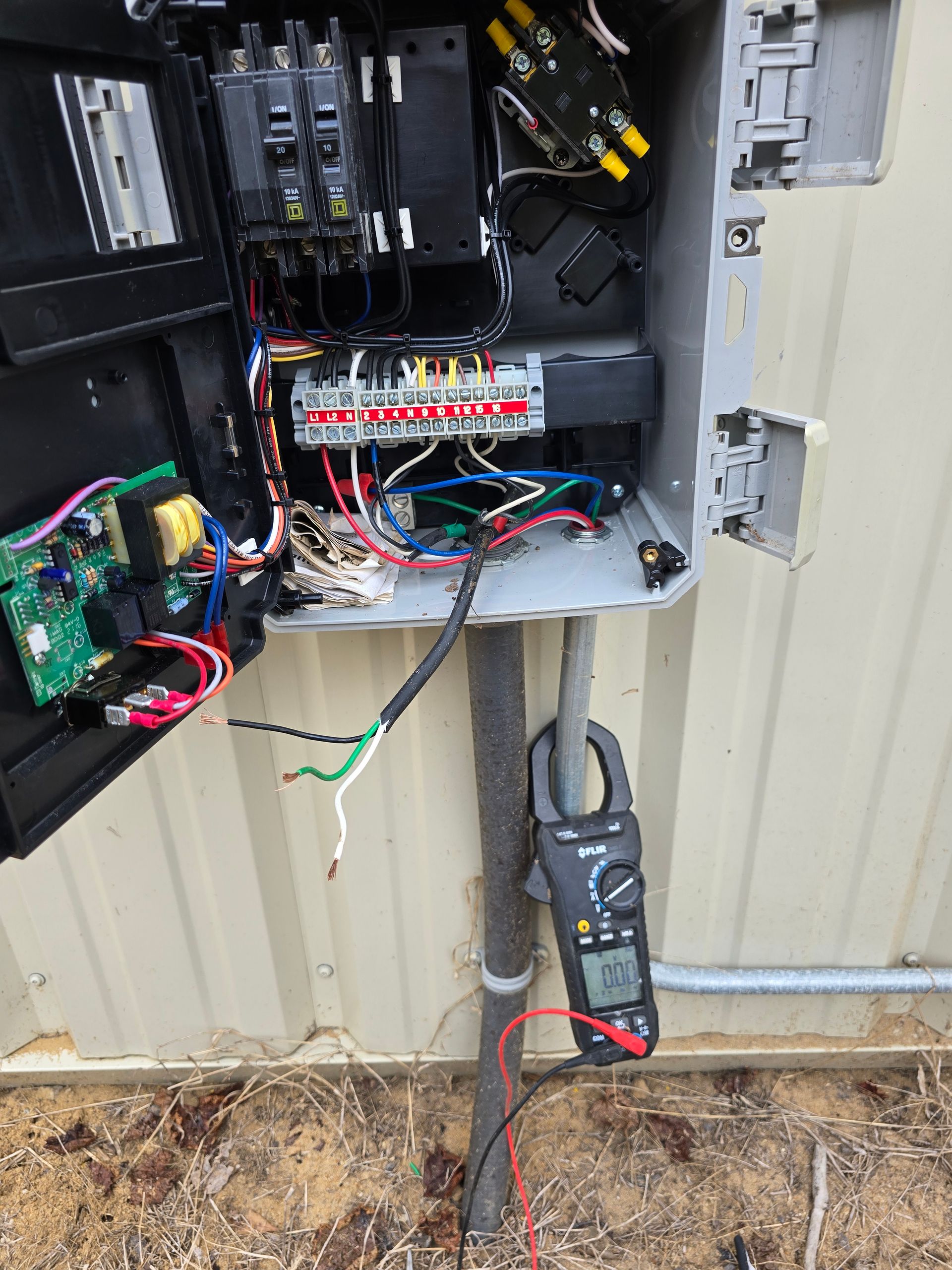 Open electrical box with wires, breakers, and a multimeter clamped to a wire, outdoors.