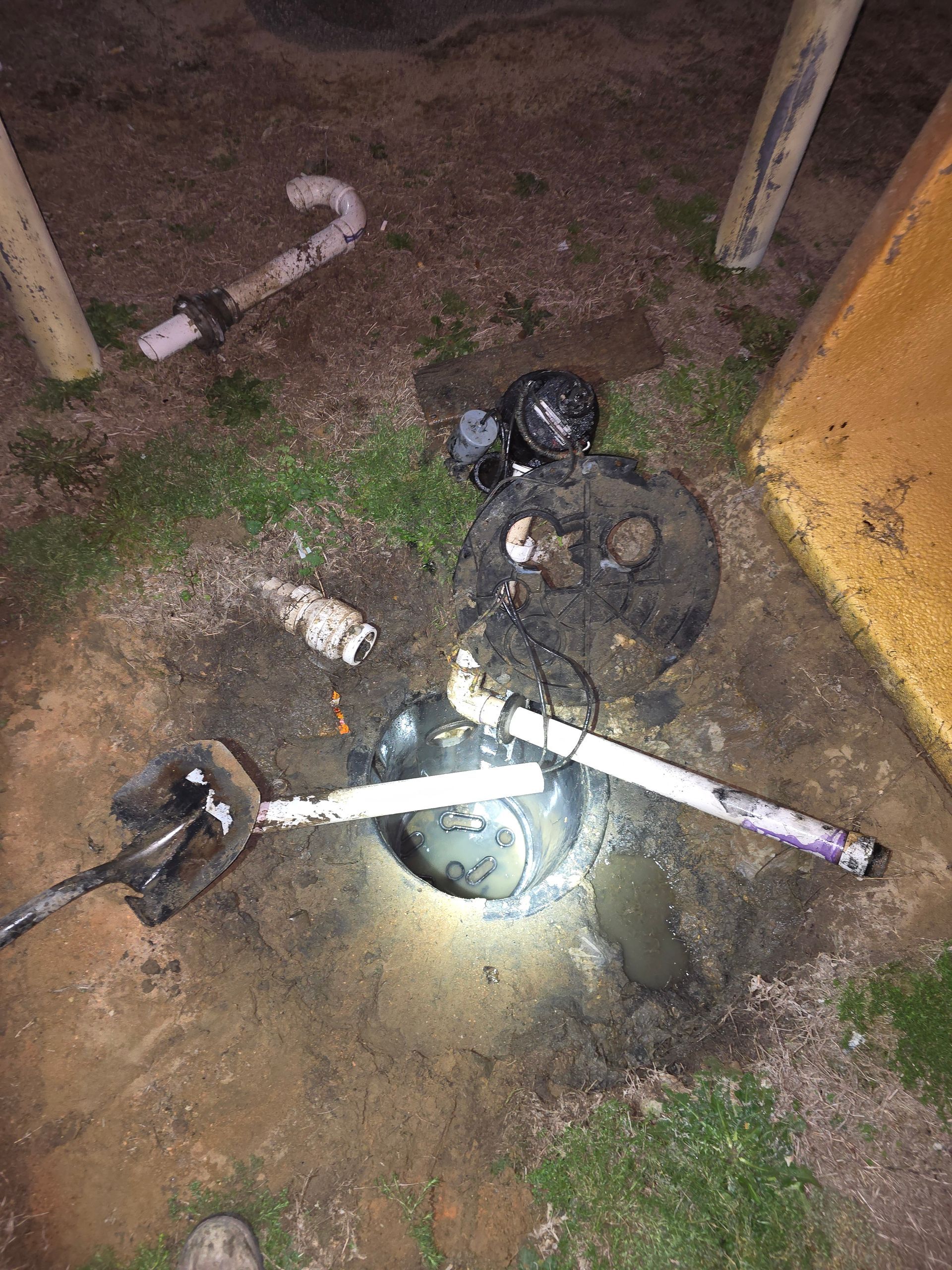 A damaged sump pump system in the ground, with exposed pipes and wires. Dark soil and standing water are visible.