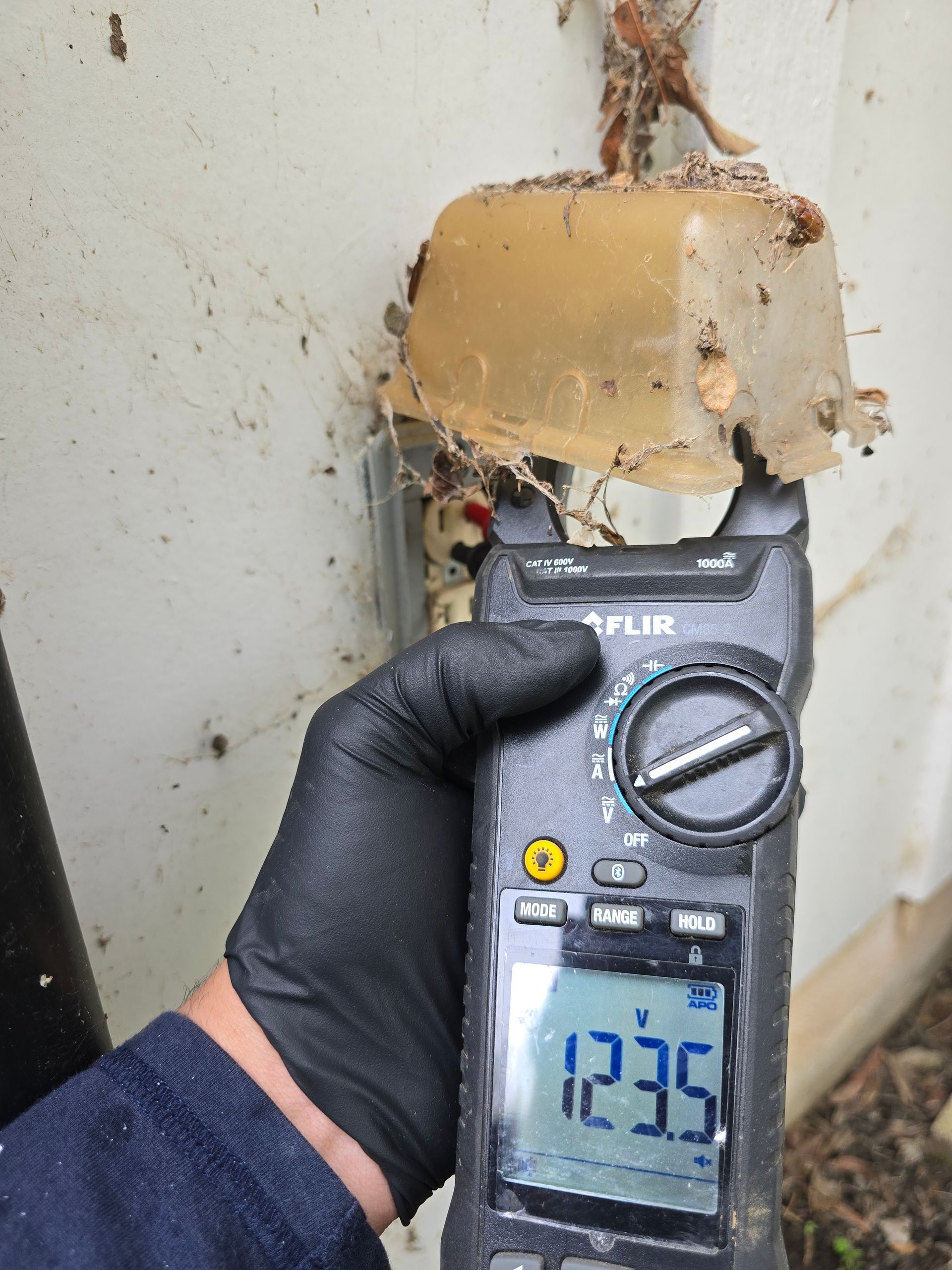 Gloved hand holding a multimeter showing 123.5 on a utility box, with a melted plastic cover above.
