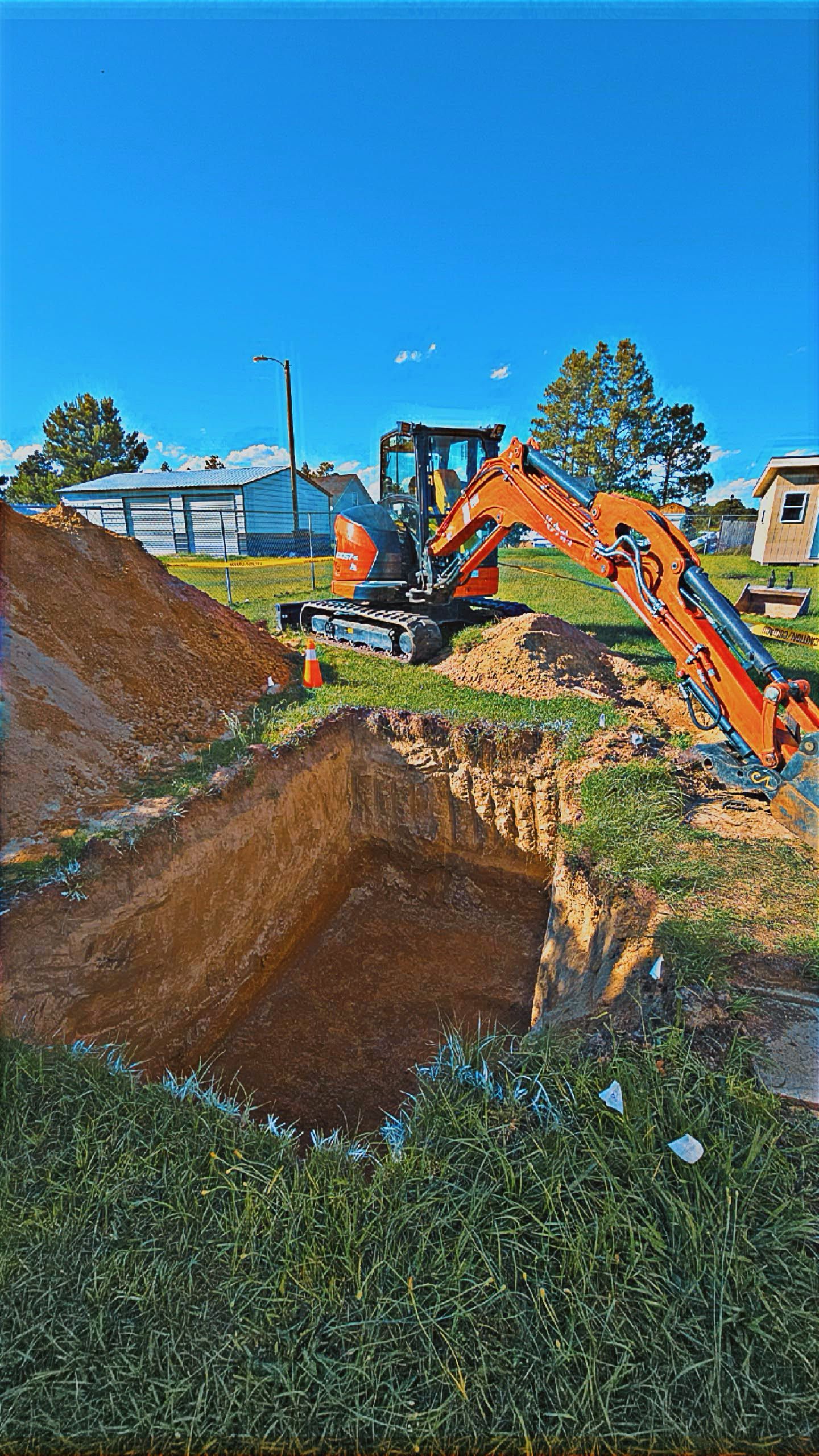 An orange excavator digs a hole in a grassy area with a dirt pile. Bright blue sky.