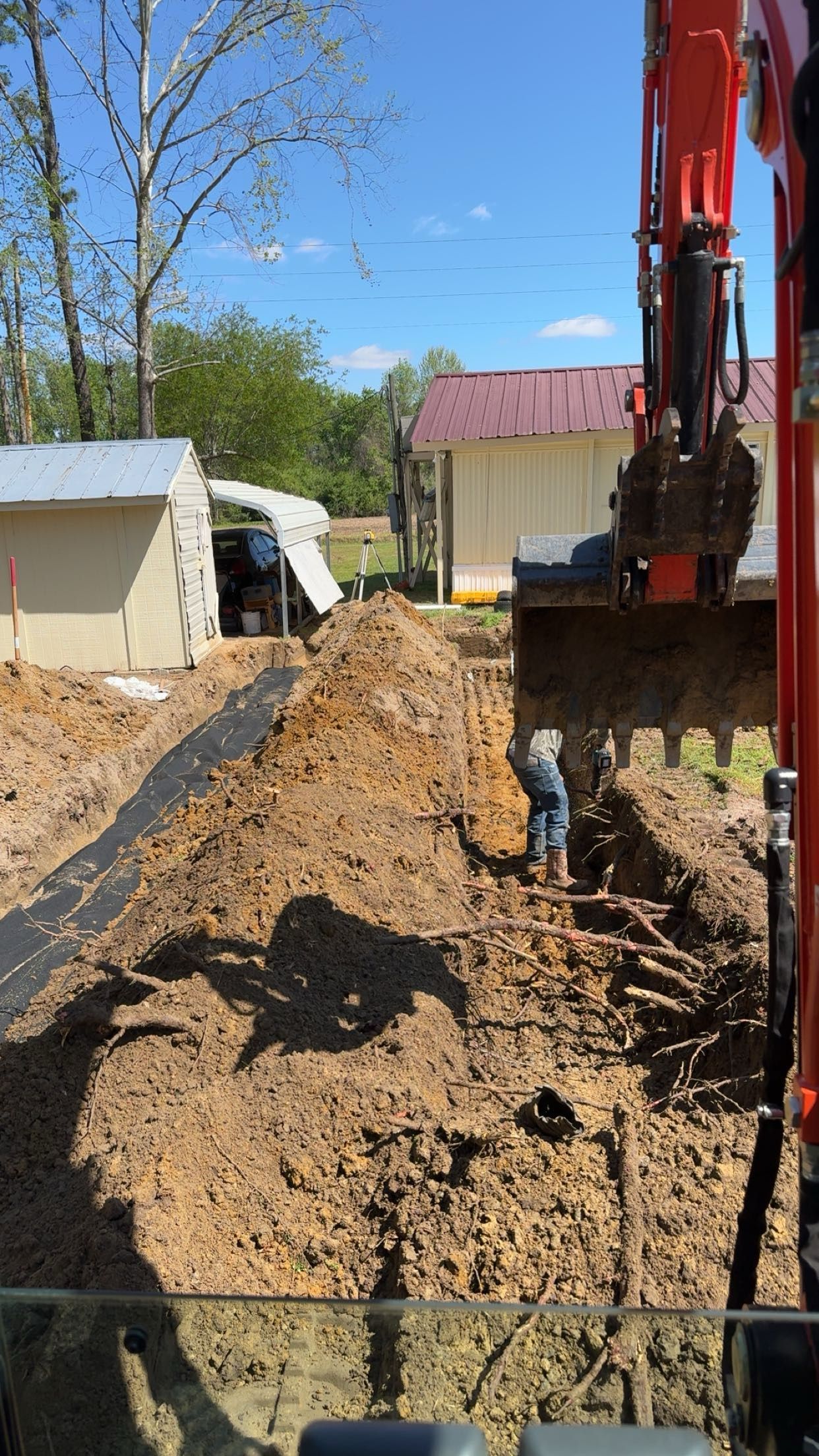 Excavator digging a trench next to a person in a work area with buildings in the background.