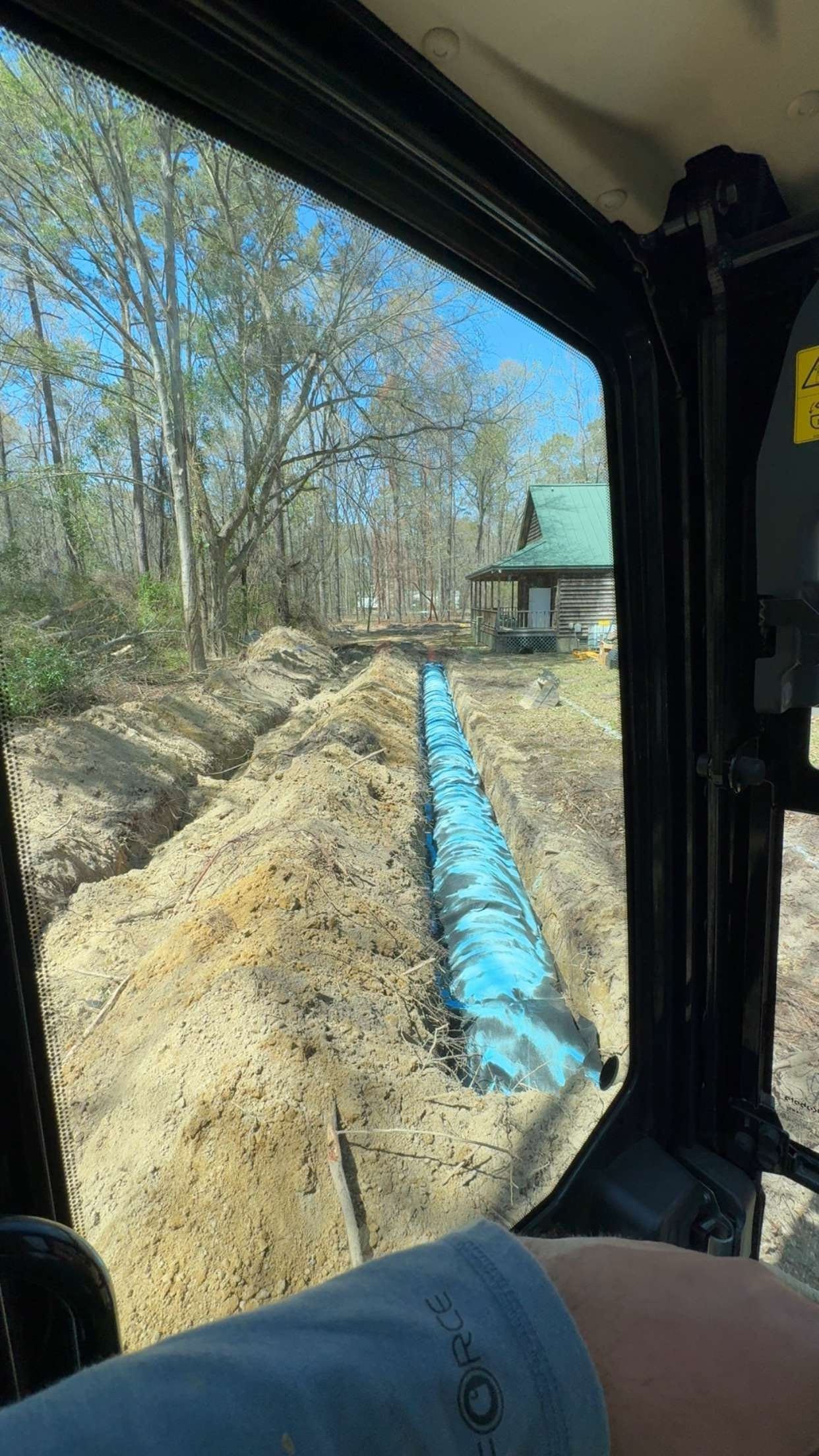 View from machinery cab over a trench with blue pipe, house in background.