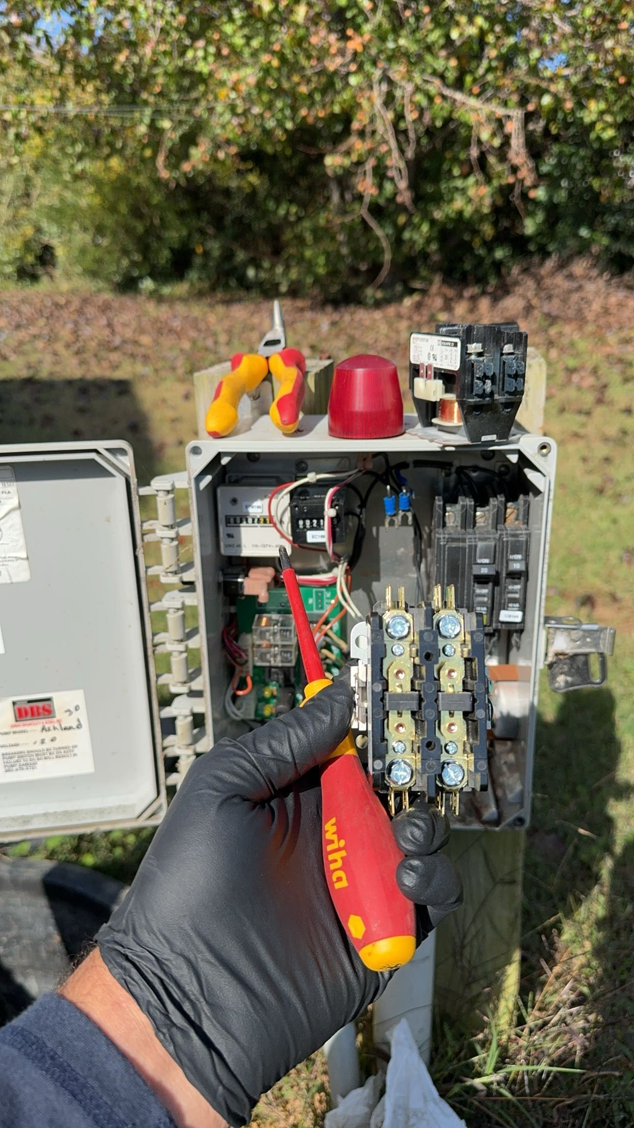 Hand in black glove using screwdriver on electrical box with exposed components, outdoors.