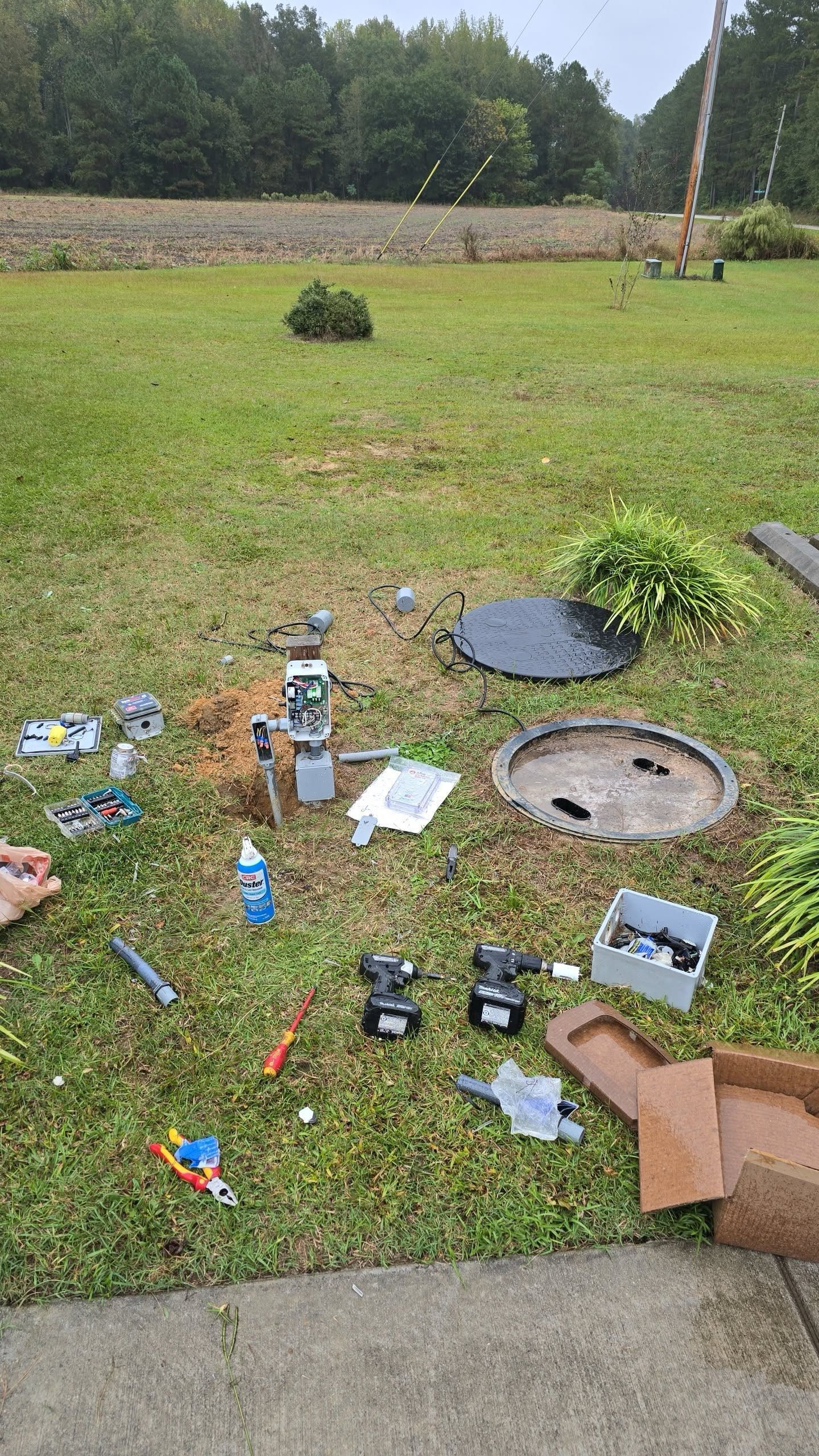 Tools and parts scattered on wet grass near two open utility covers.