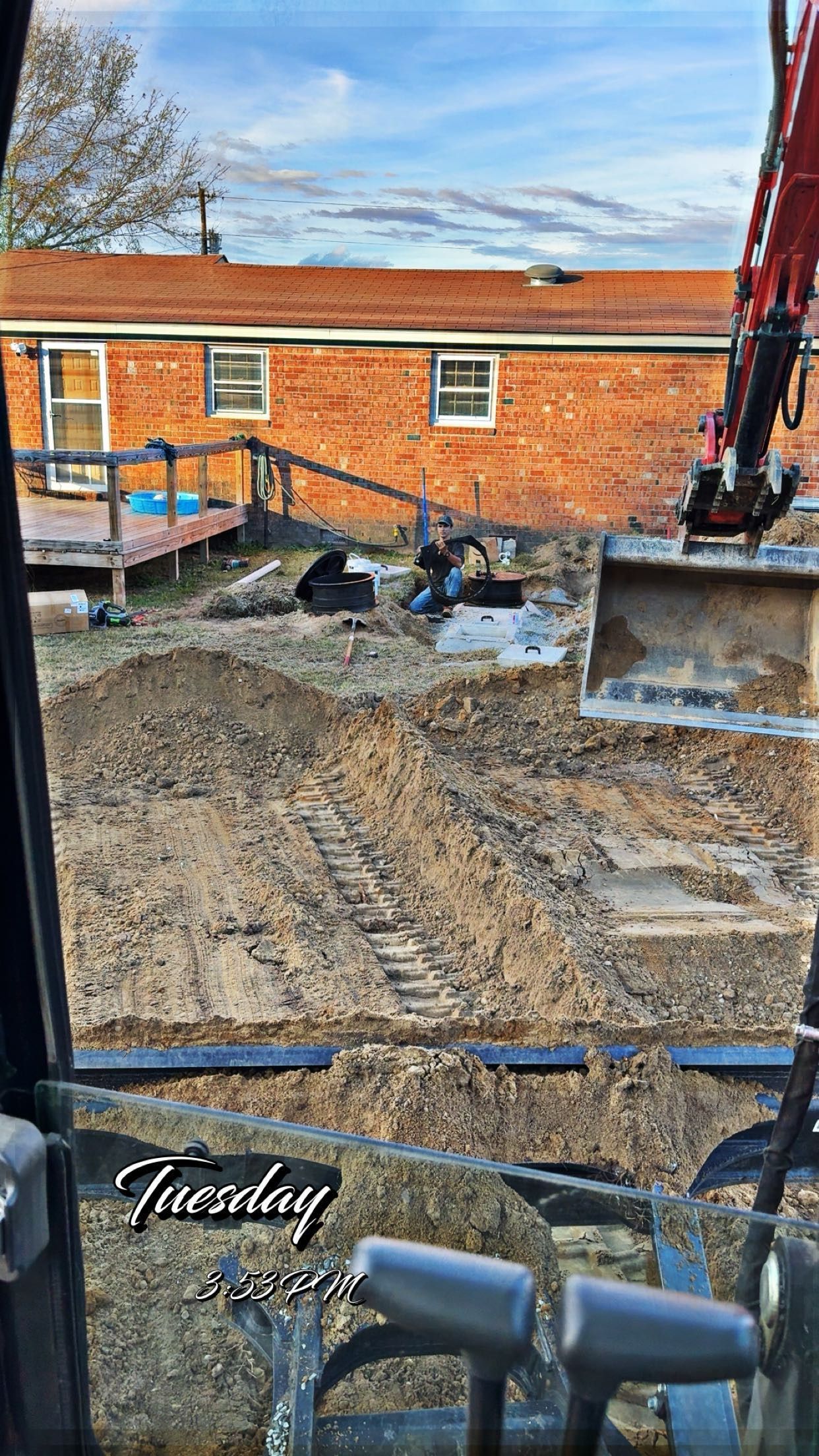 Excavator in backyard, digging into dirt, with a brick house and blue sky in background. 