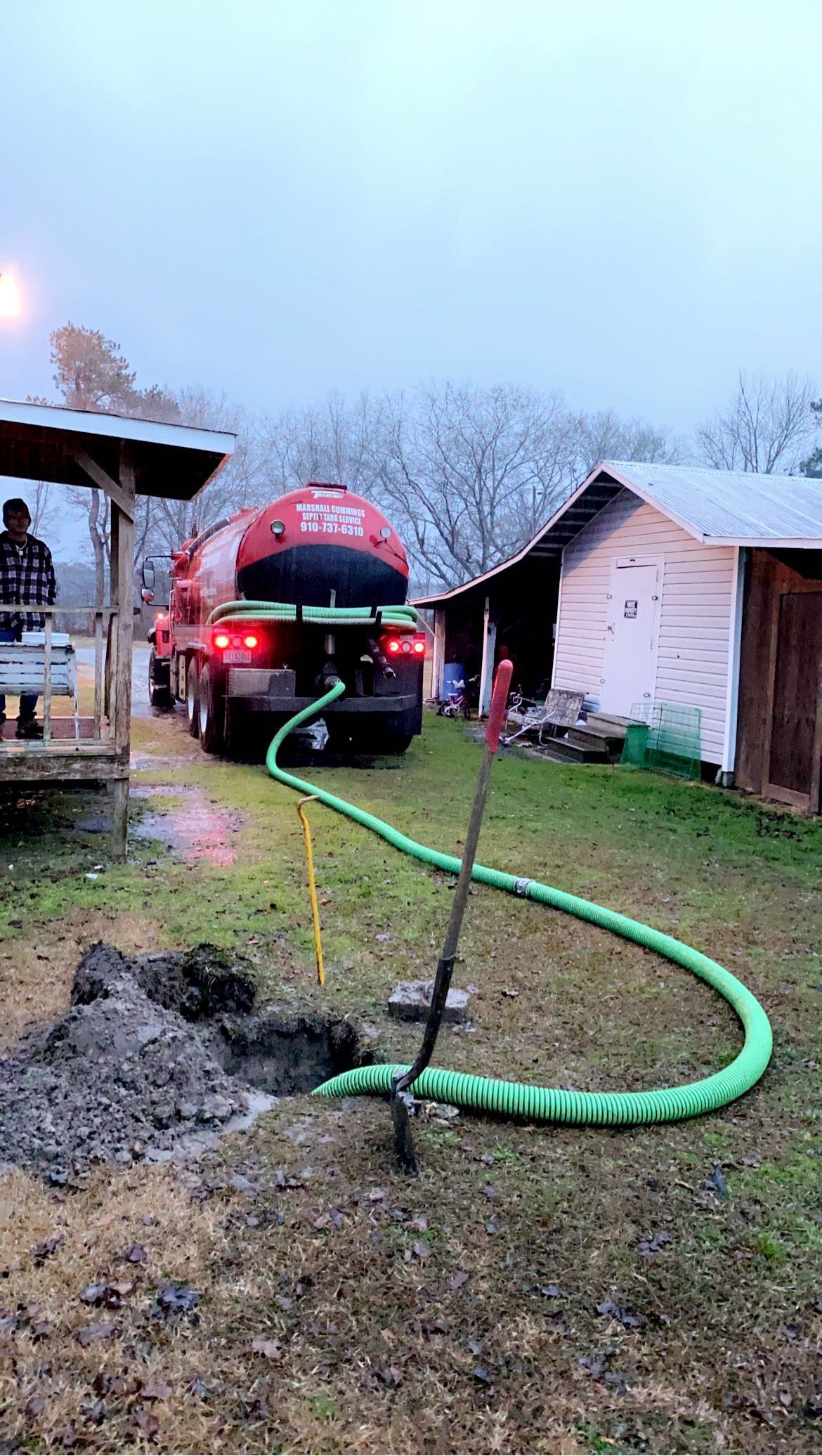 Septic tank truck emptying a septic system. A green hose extends from the truck to a muddy hole. Cloudy sky and snowy trees in background.