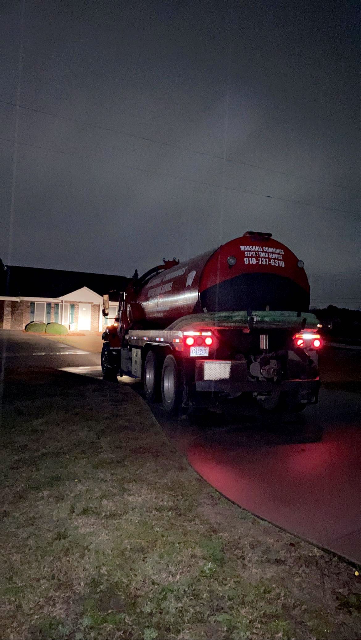 Tanker truck parked in front of a house at night, illuminated by red taillights and porch light.