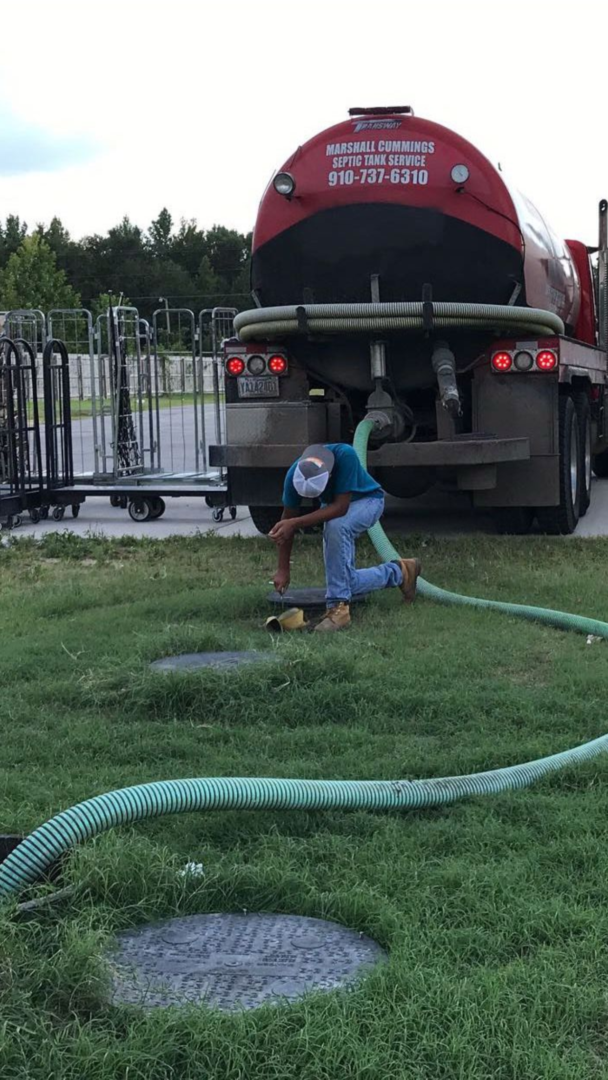 A worker connects a green hose to a septic tank, near a red tanker truck on a grassy area.
