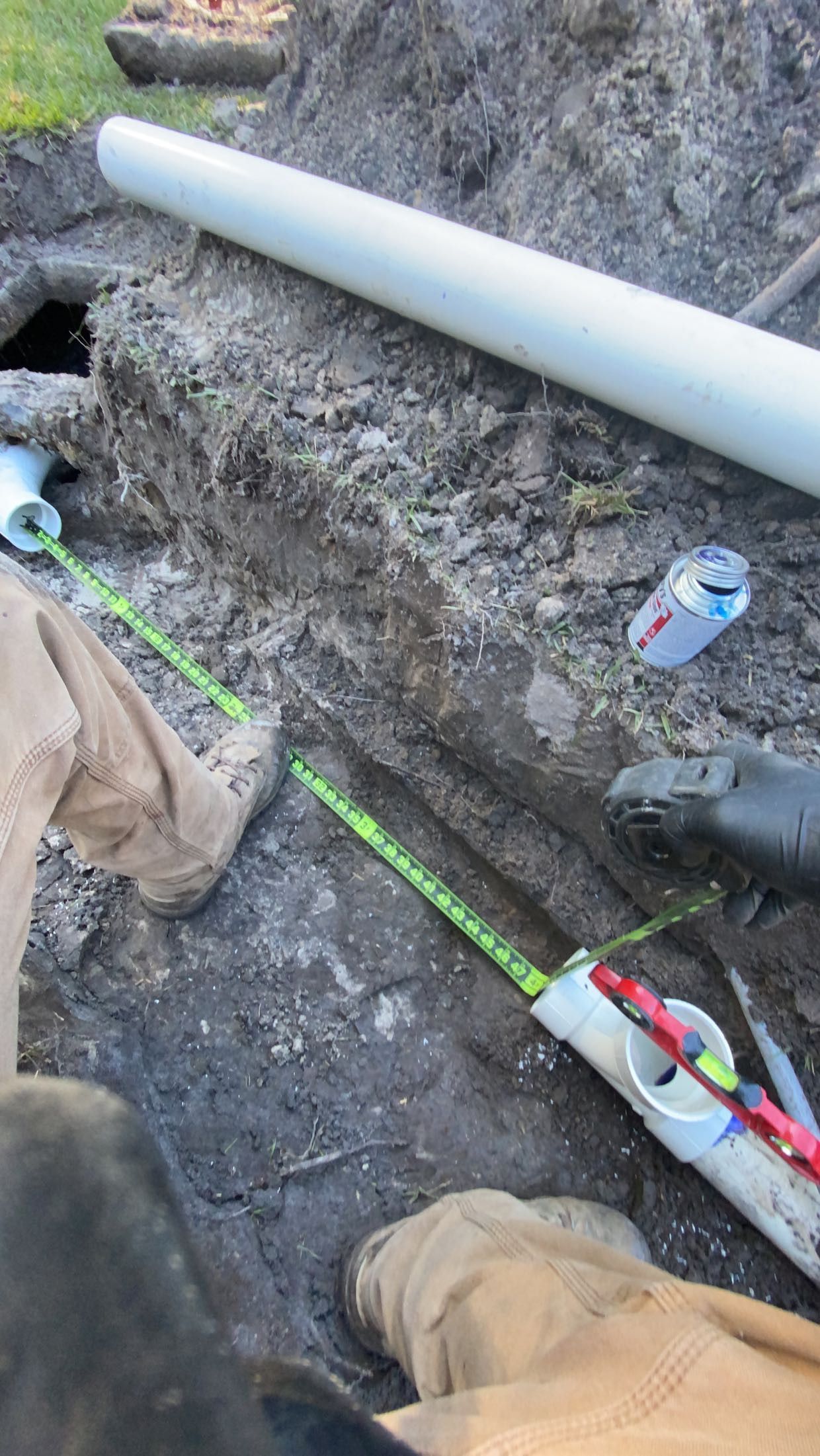 Trench with white PVC pipes, a can, and a worker's boots. Green and red wires are visible within the pipes.