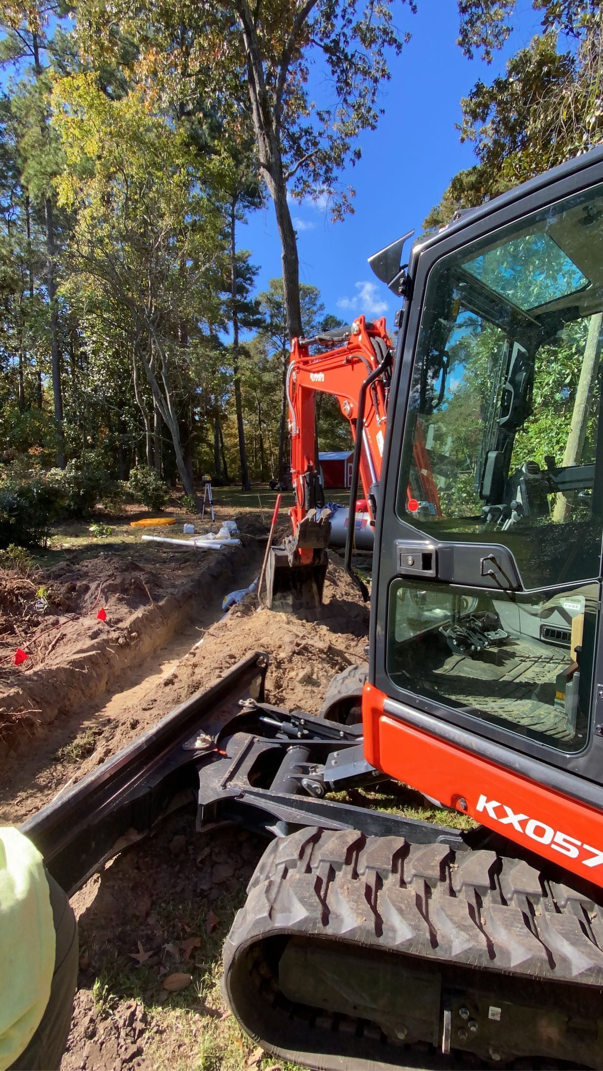 Orange excavator digging a trench in a yard, trees in the background, blue sky.