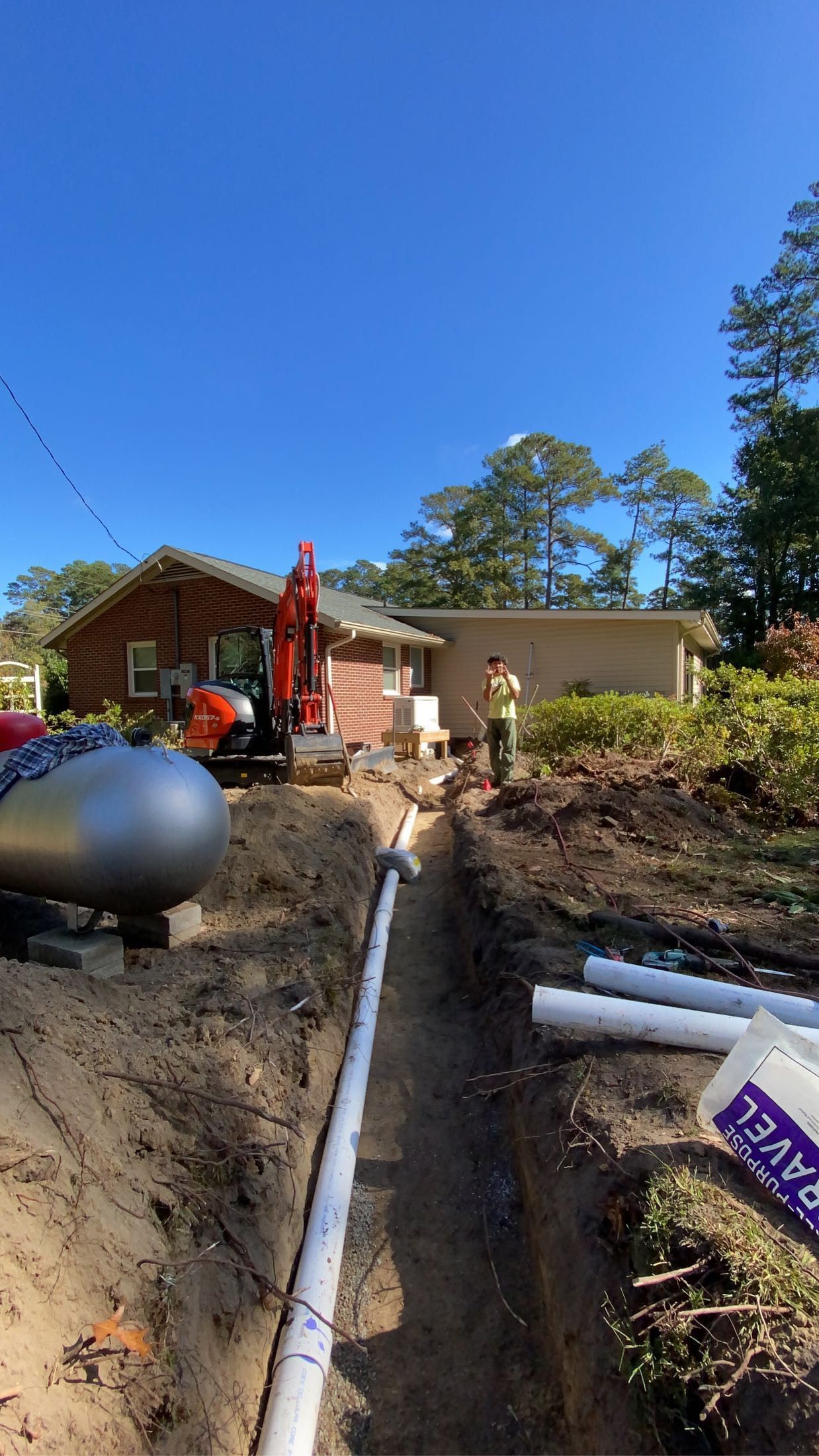 Construction site with an excavator digging a trench for a white pipe near a house under a blue sky.