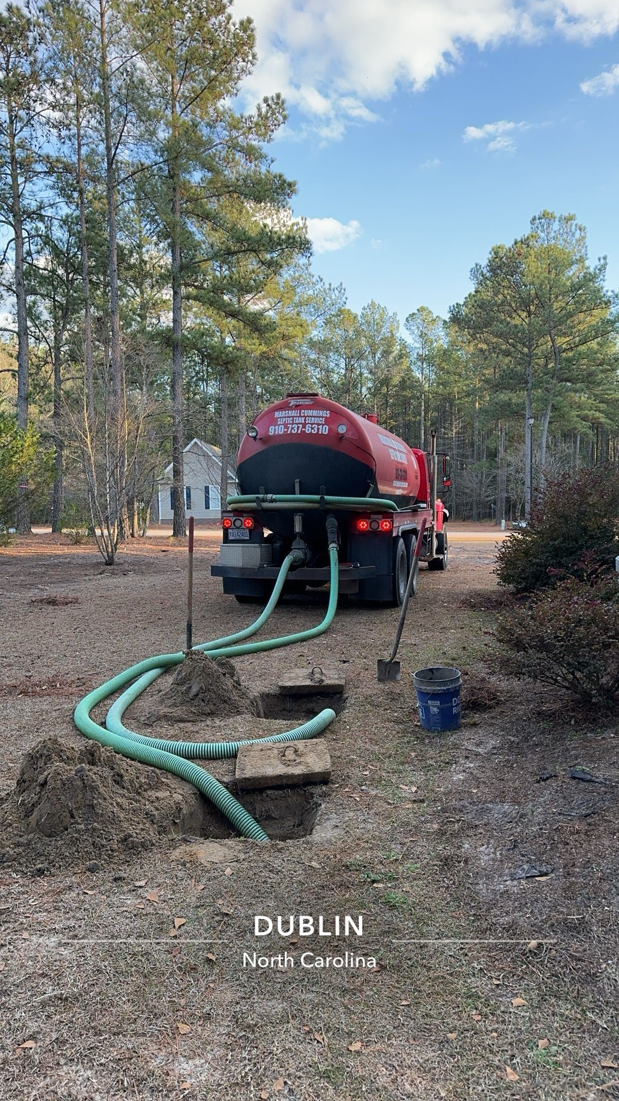 Red septic truck with hoses in holes in a yard, trees in background, blue sky.