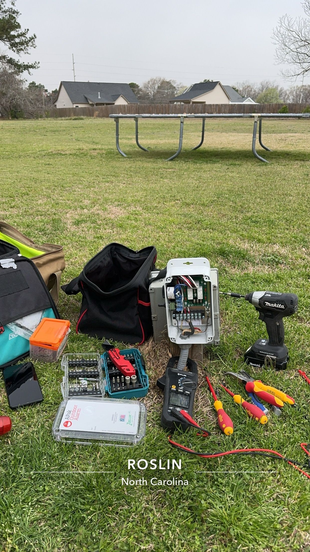 Tools and disassembled device on grass, outdoor setting. Screwdrivers, drill, black bag, and toolbox.