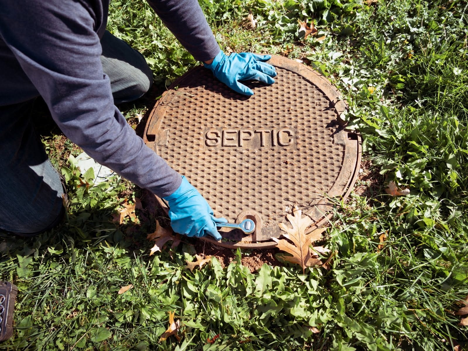 Person in blue gloves opening a septic tank lid in grassy area.