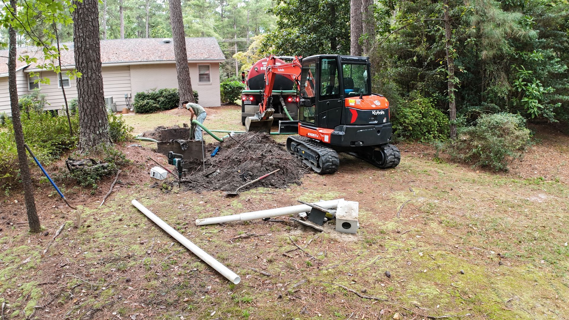 Orange and black trackhoe working near a septic tank in a yard with trees and a house.