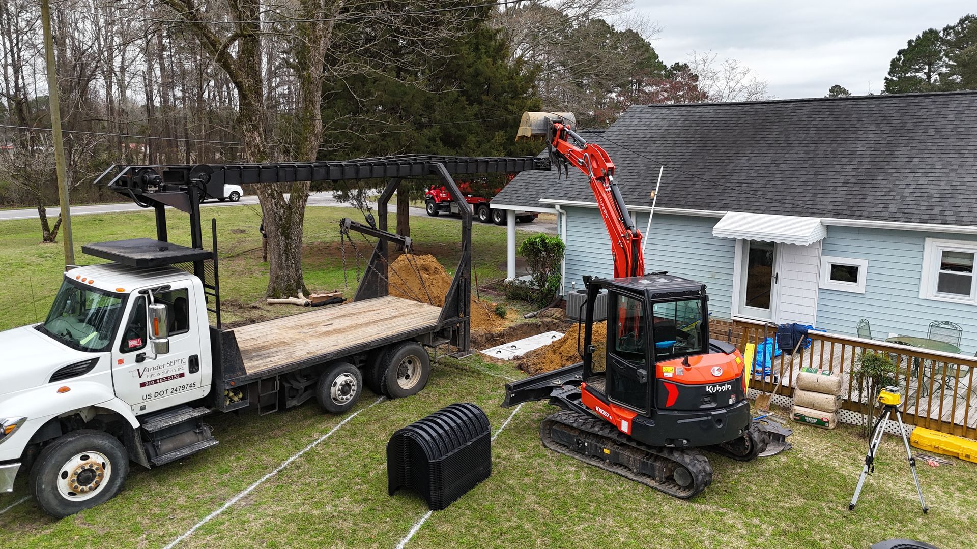 An excavator digging near a house, with a flatbed truck and a dark container, outdoors.