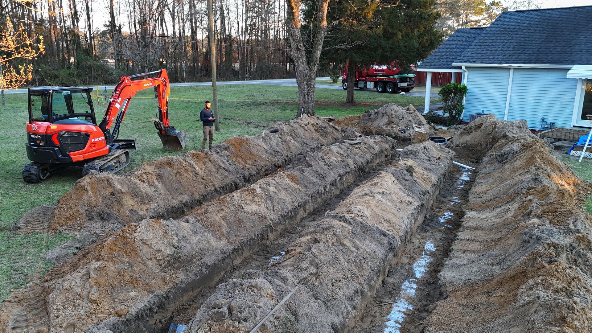 Excavator digging trenches in a yard; a worker stands nearby. Blue house and trees in the background.