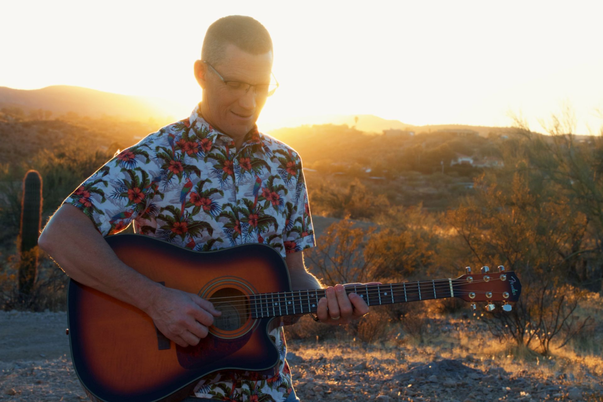 Man in a tropical shirt playing guitar at sunset, surrounded by desert scenery and cacti.