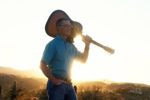 A man in a blue shirt is holding a guitar in front of a sunset.