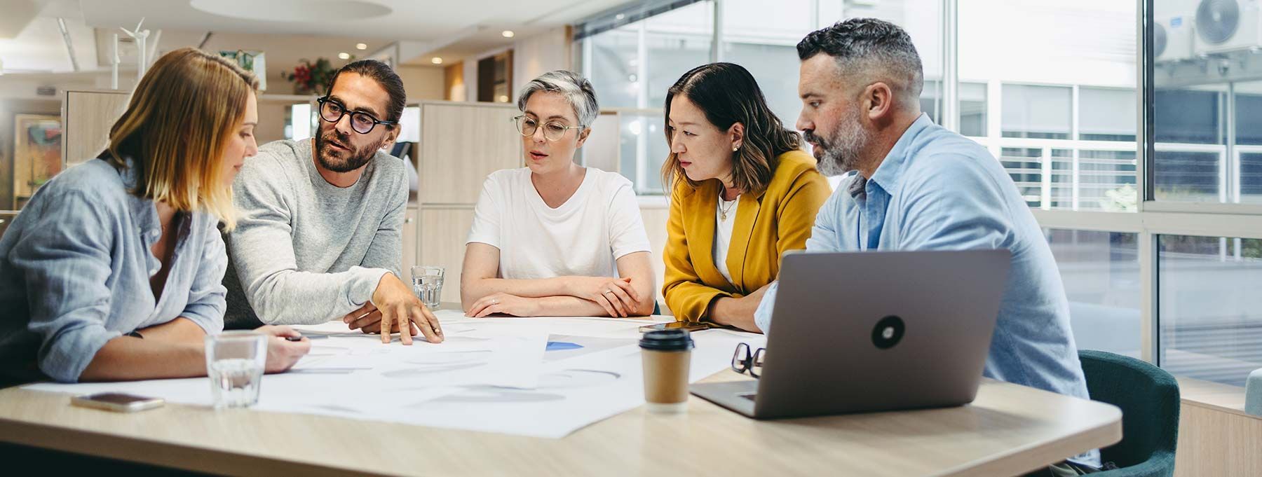 Group of coworkers in a bright office meeting around a table with papers, laptop, and coffee cup