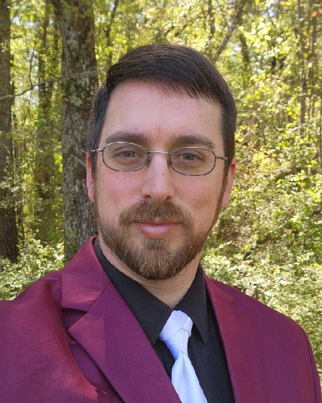 Portrait of a man in a burgundy jacket and white tie, standing outdoors in a wooded area.