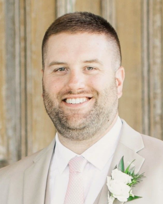 Smiling man in a beige suit with a white boutonniere and pink tie, standing in front of wooden doors