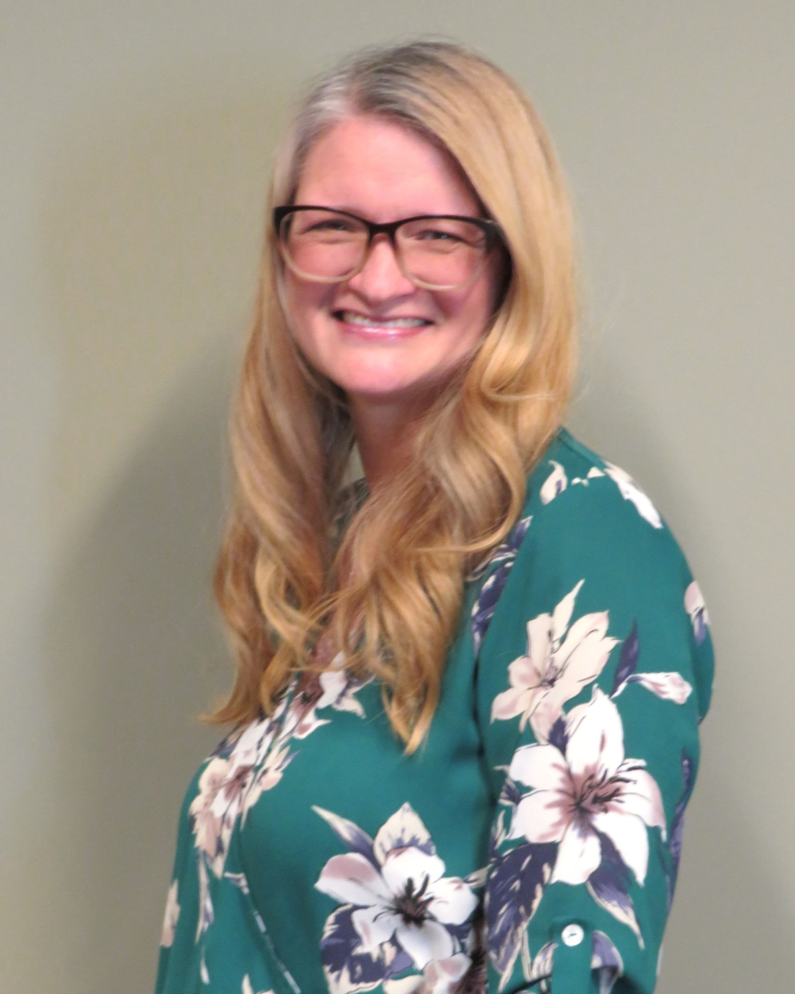 Smiling person with glasses in a green floral blouse against a plain wall