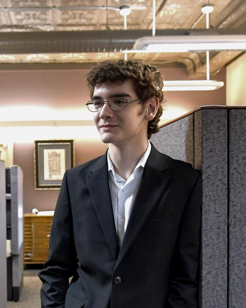 Man in a dark suit and glasses standing in an office, looking to the side.