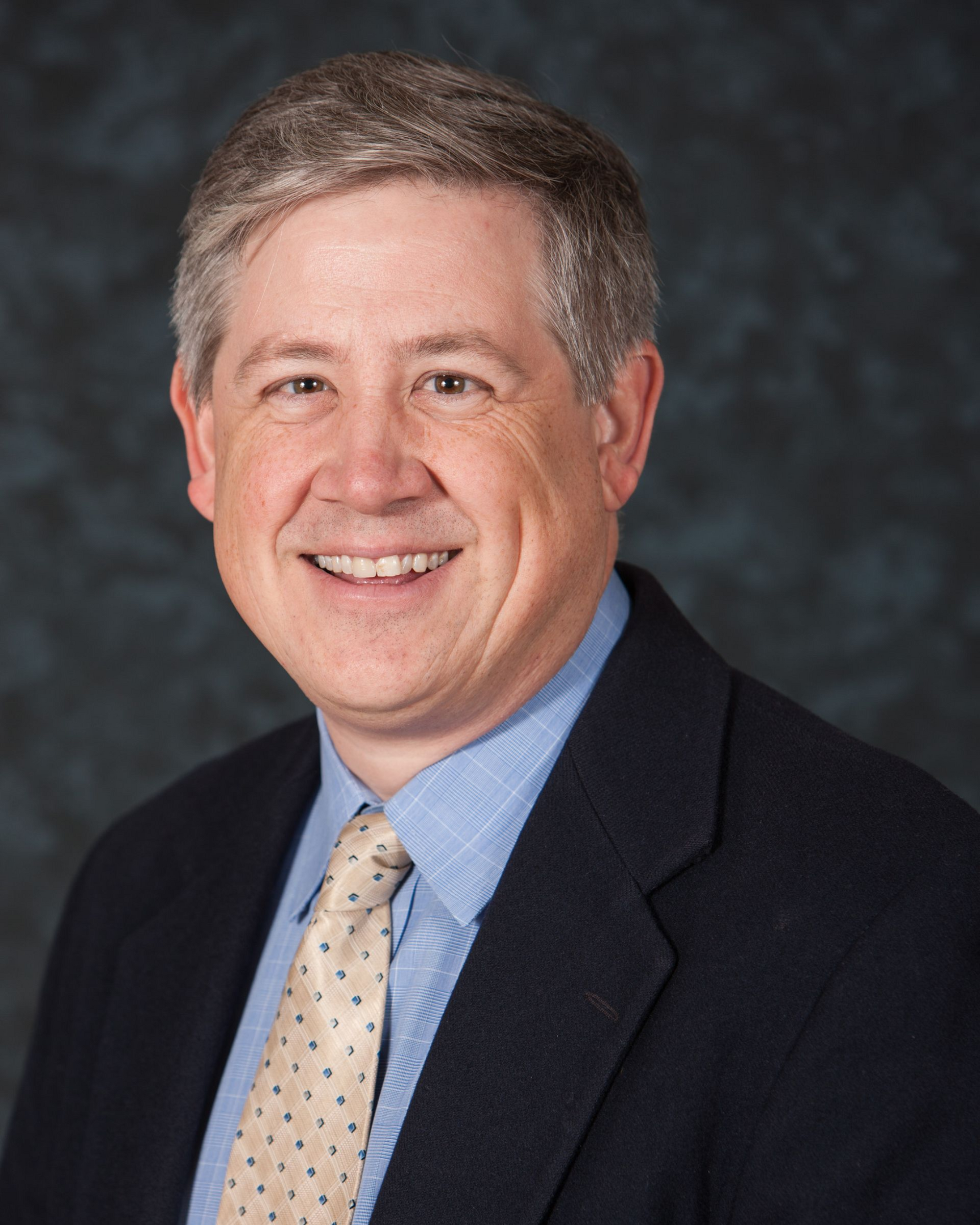 Professional headshot of a man in a dark suit, blue shirt, and patterned tie against a gray backdrop