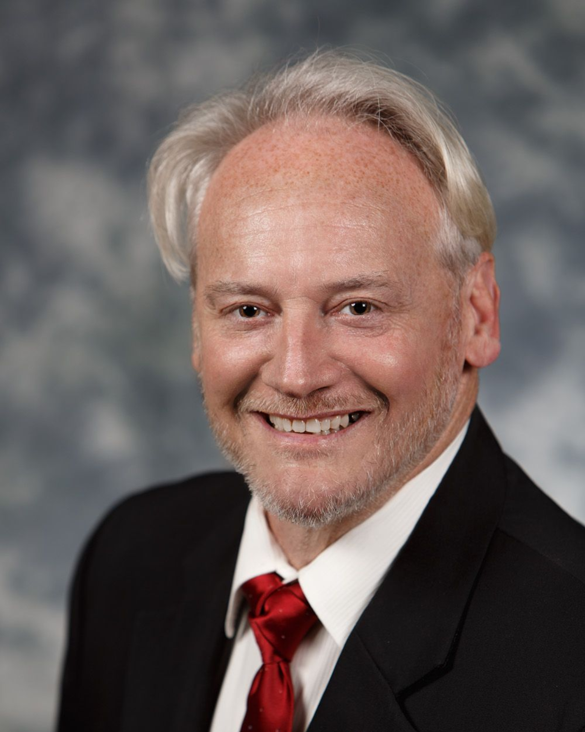 Studio headshot of a smiling person in a black suit and red tie against a gray backdrop