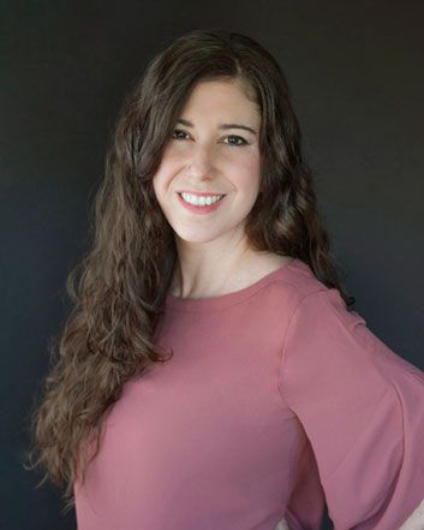 Smiling person with long wavy hair in a pink top against a dark background
