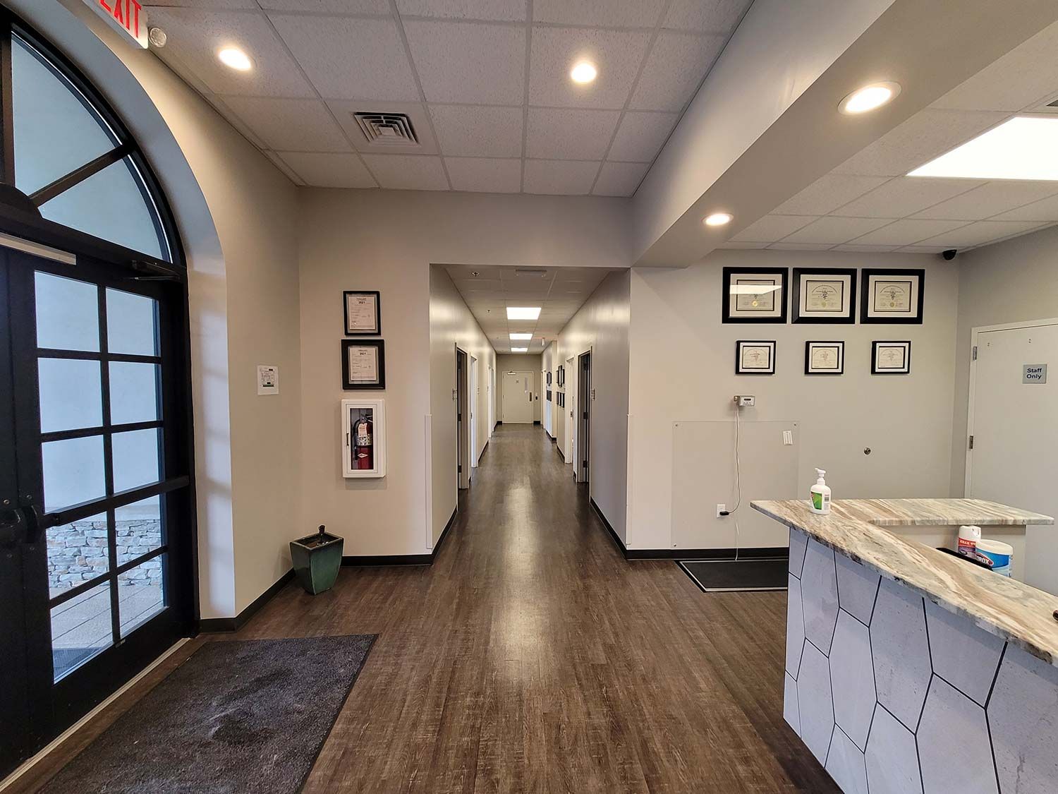 Bright office hallway with framed certificates, reception counter, and arched window; long corridor recedes ahead.