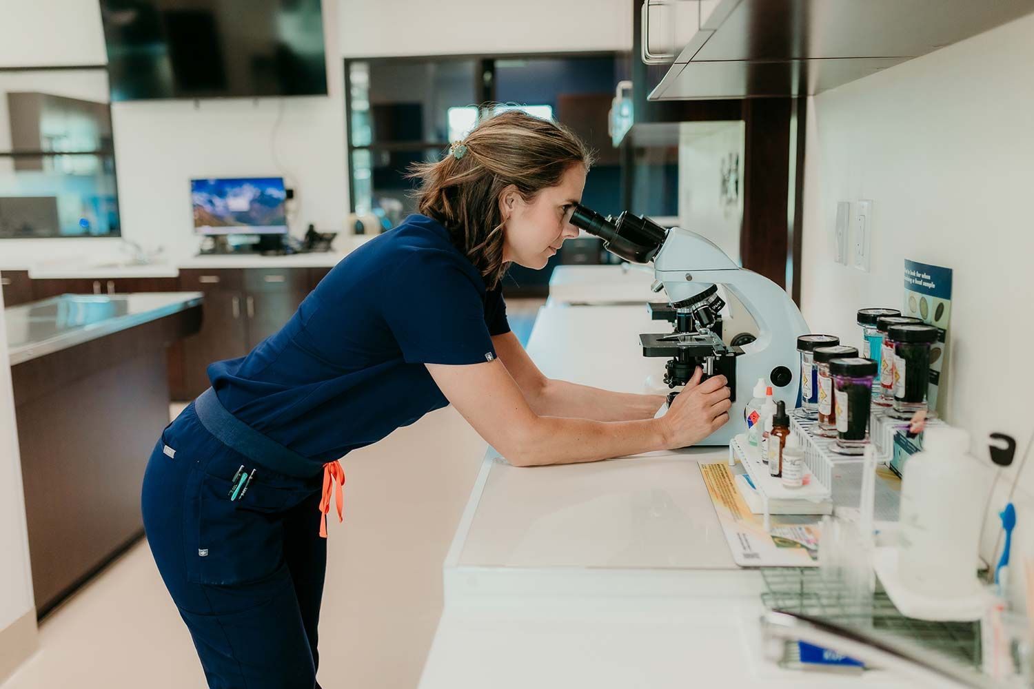 Person in a blue shirt leaning over a lab bench, examining a microscope in a bright lab