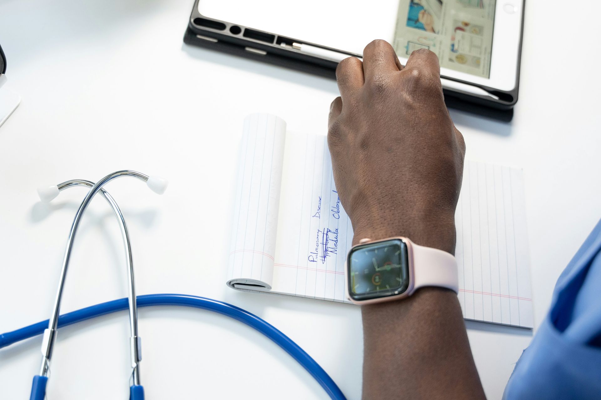 A person wearing an apple watch is writing on a notepad next to a stethoscope.
