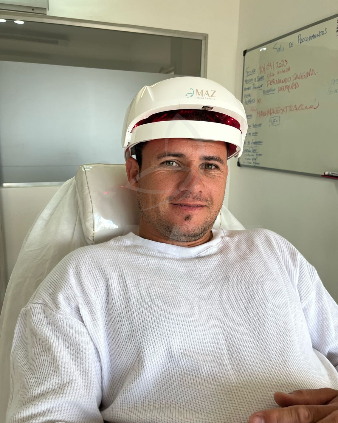 Paciente sonriente tras implante capilar, con venda postquirúrgica y bata en sala de recuperación.