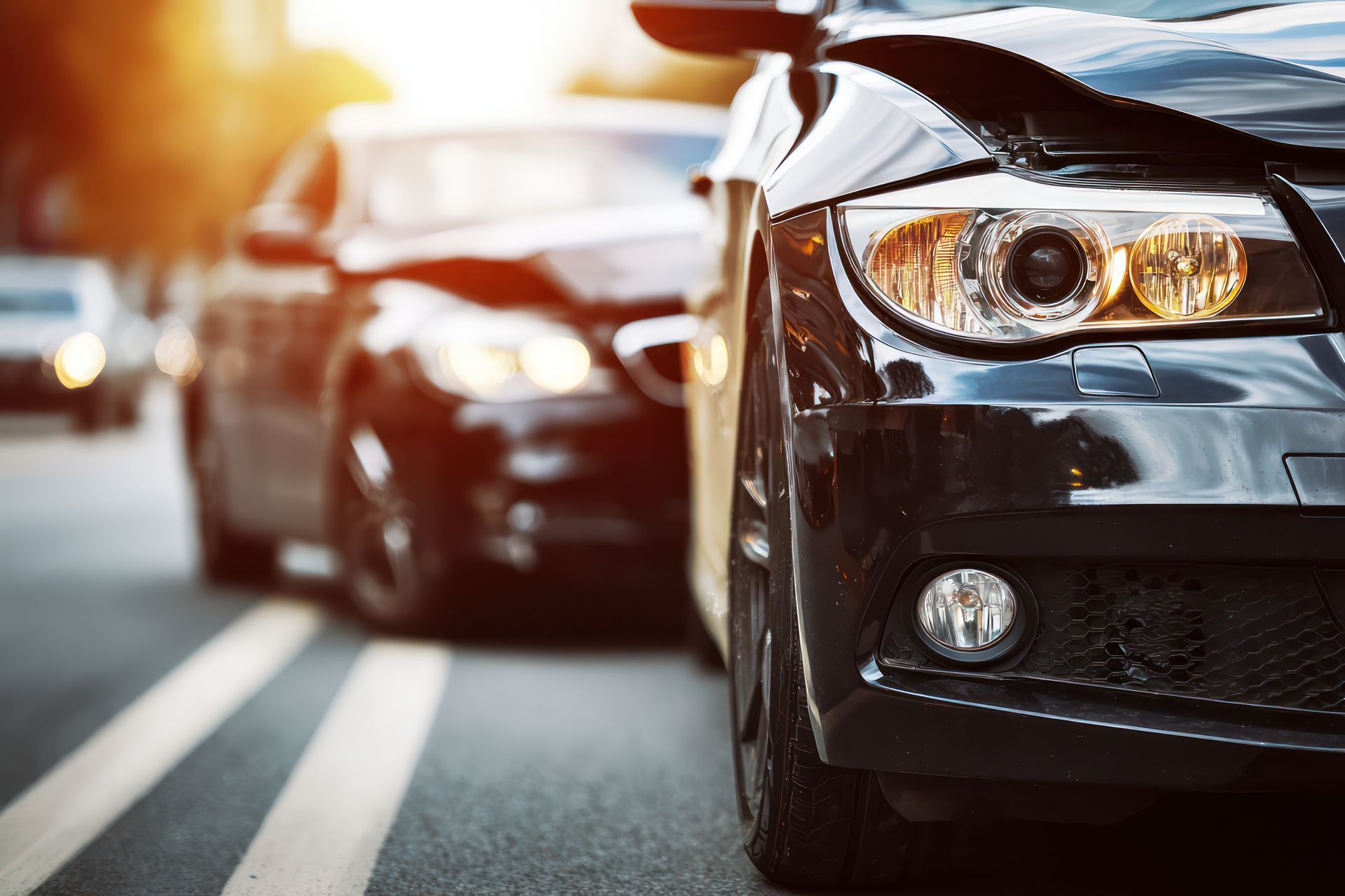 Close-up view of the front of a damaged black car, with another car partially visible in the background on a road.