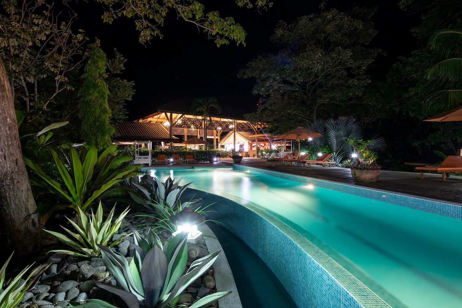 A large swimming pool is lit up at night with a house in the background.