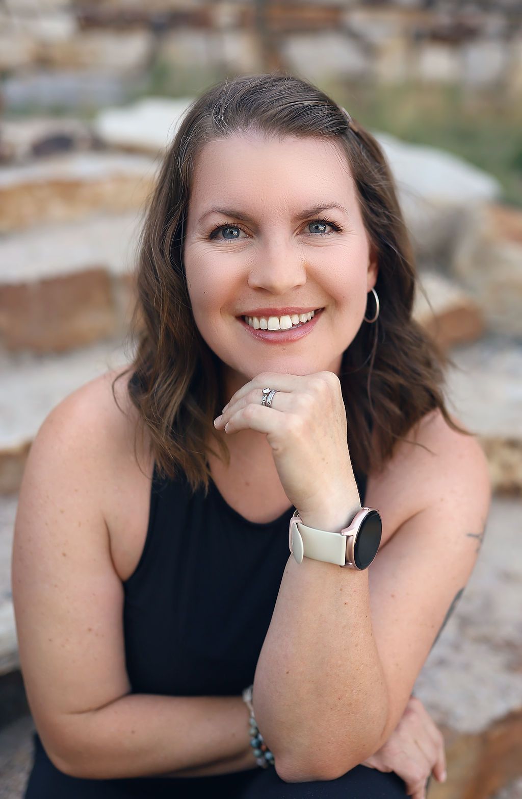 A woman wearing a black tank top and a watch is smiling with her hand on her chin.