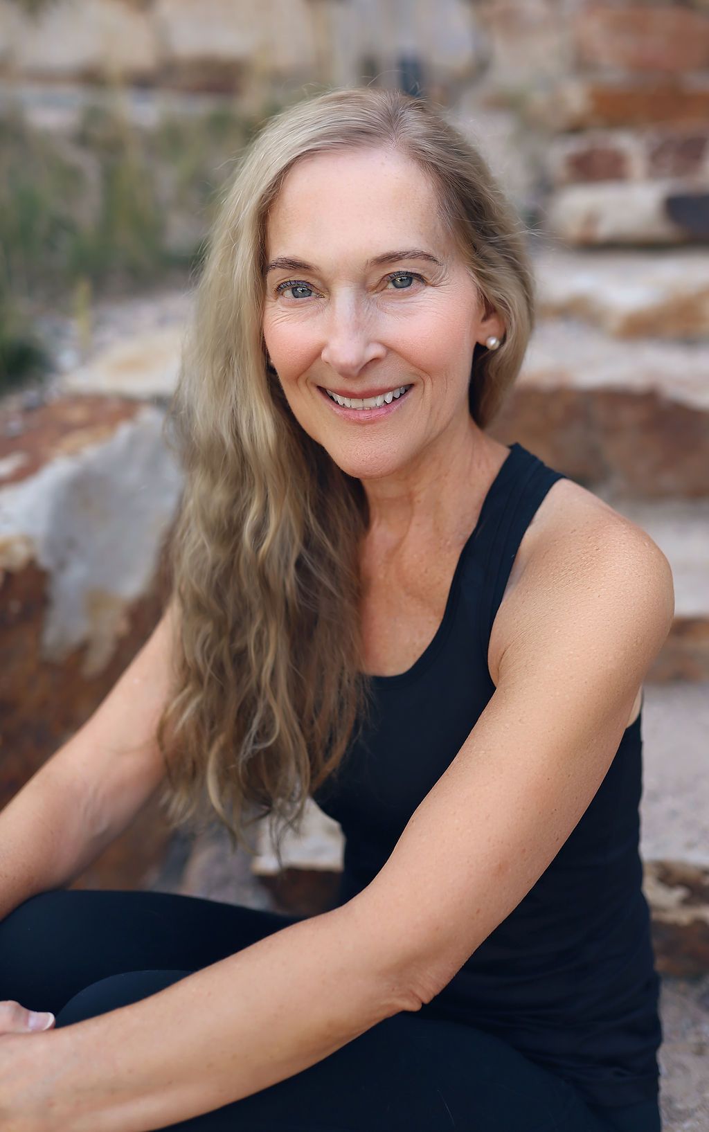 A woman in a black tank top is sitting on a rock and smiling.