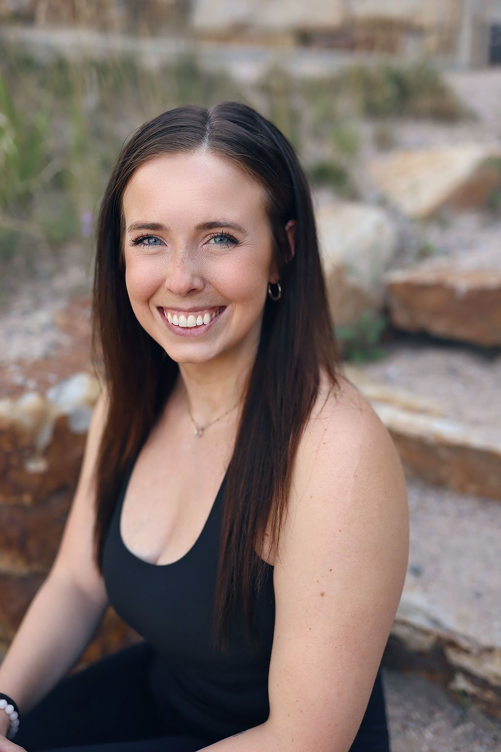 A woman in a black tank top is smiling while sitting on a rock.