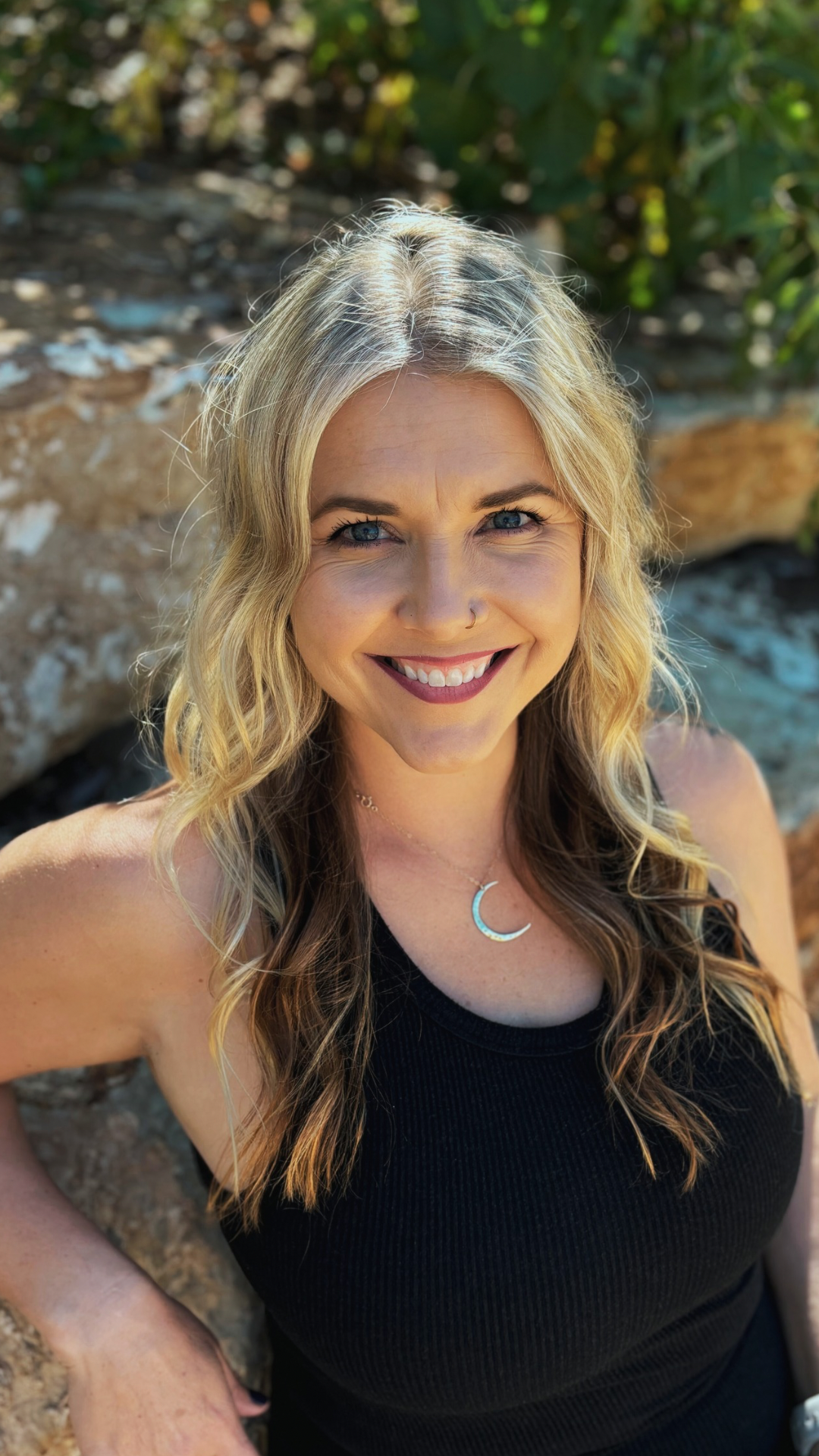 A woman in a black tank top is smiling for the camera while sitting on a rock.
