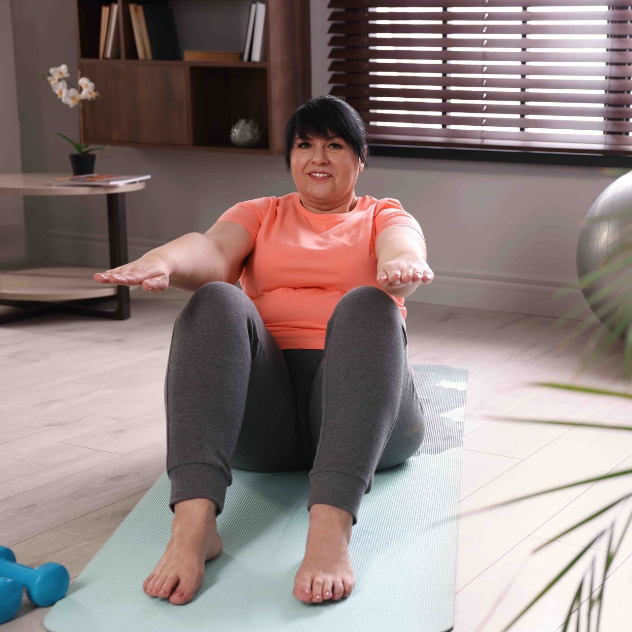 A woman is sitting on a yoga mat in a living room.