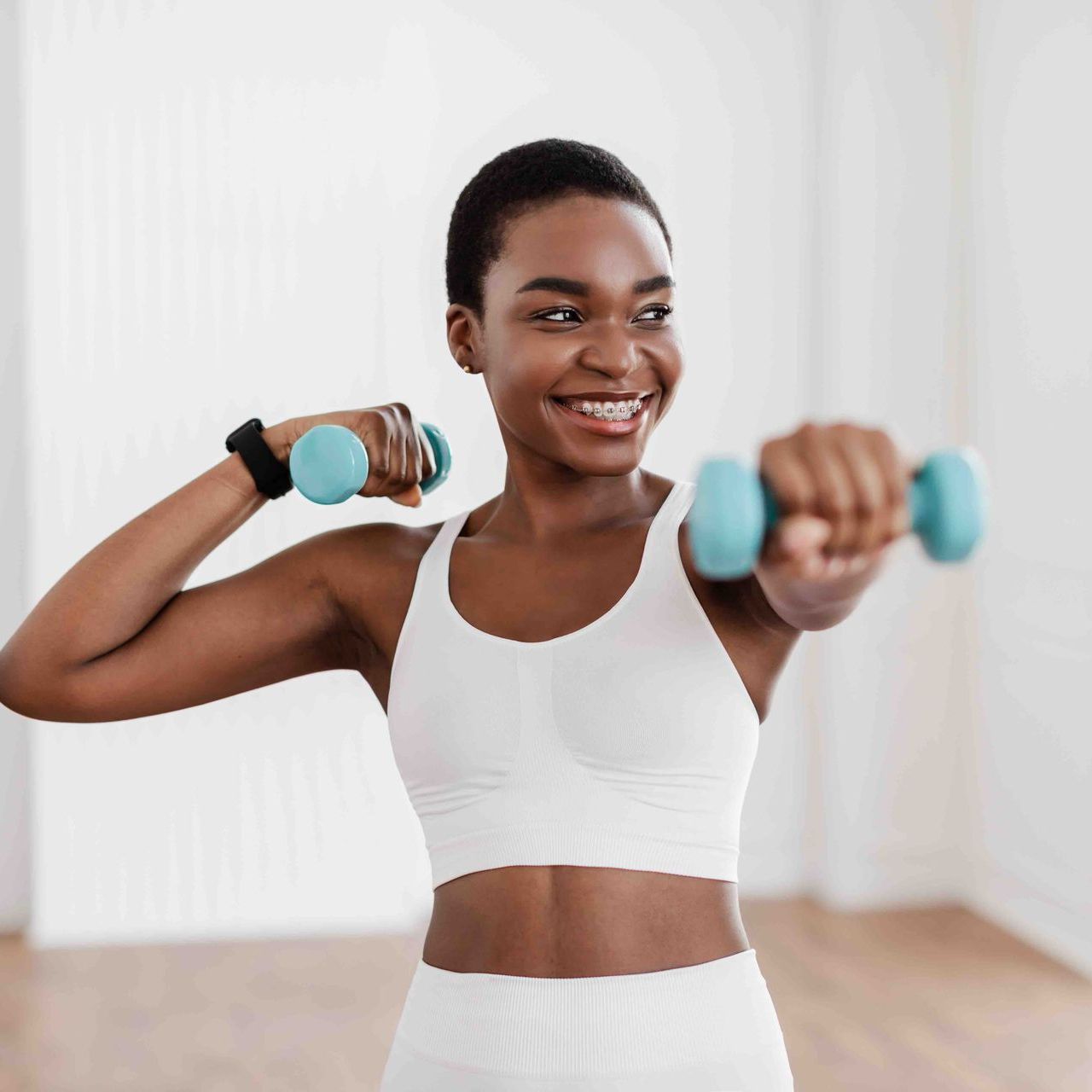 A woman is smiling while holding two blue dumbbells.