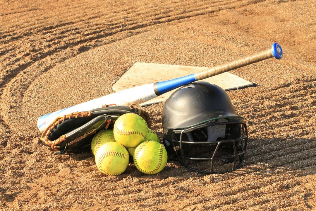 Baseball bat, helmet, glove, and three softballs on a dirt field